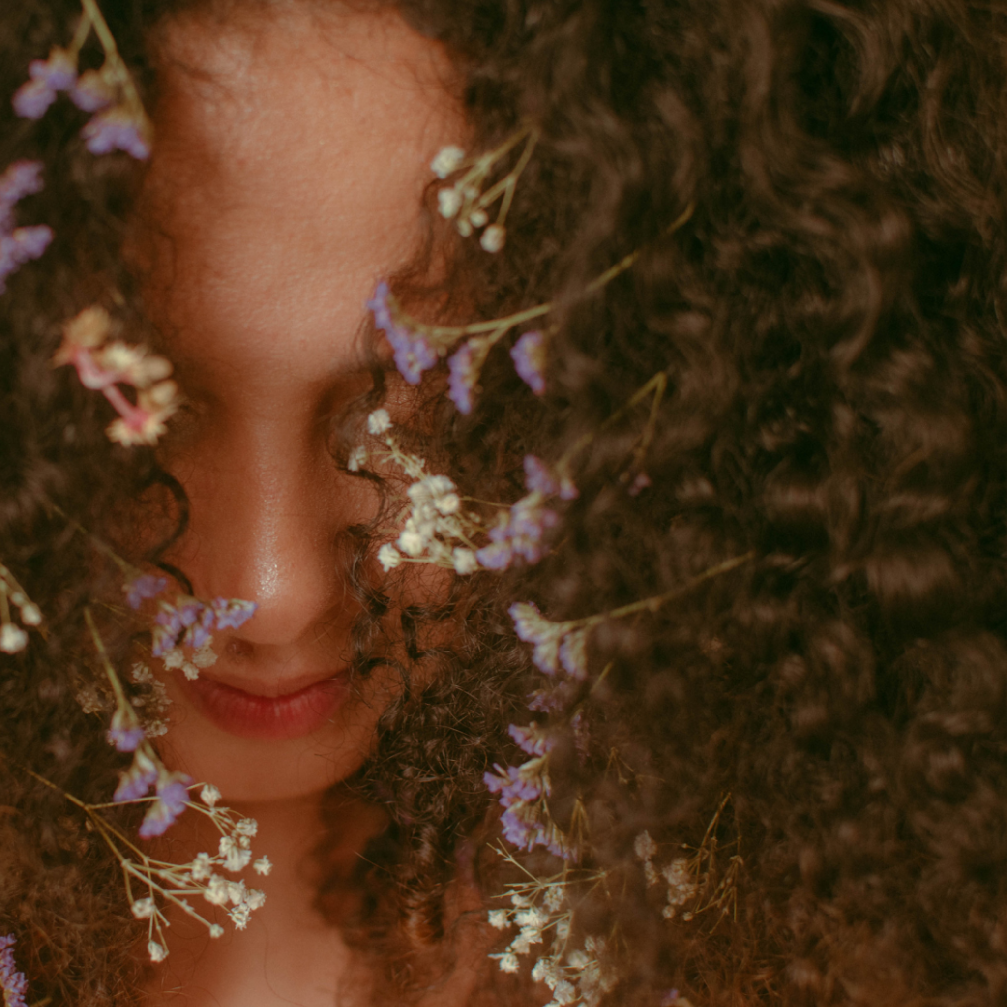 Woman with curly brown hair decorated with white and purple dried flowers.