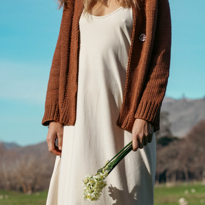 Person wearing a white dress and brown cardigan holding flowers in a field
