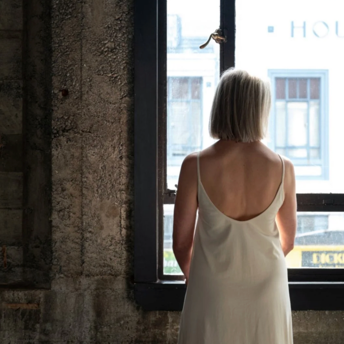 Woman in a white dress standing by a window with a blurred cityscape outside.