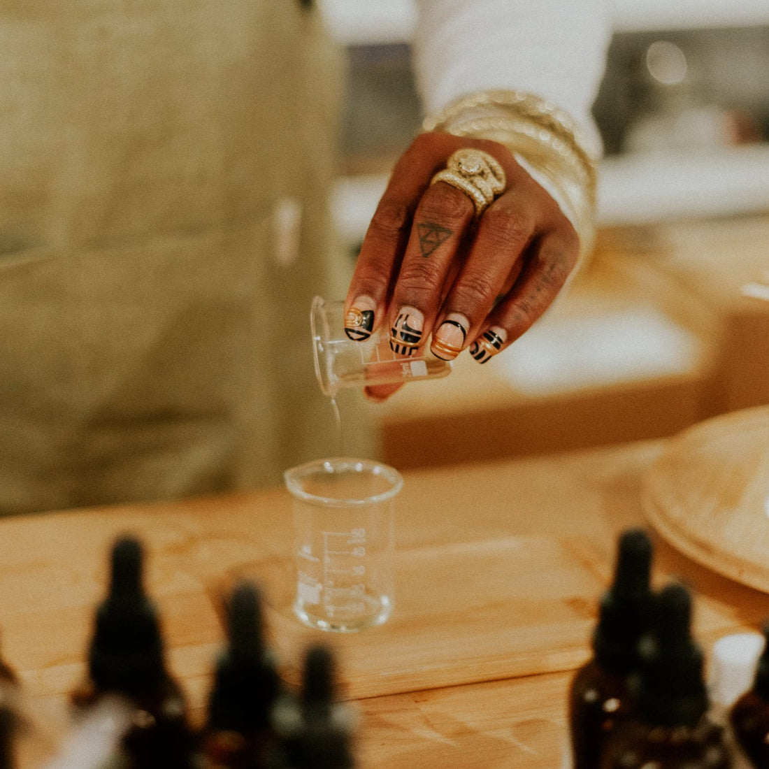 Hand with intricate nail art holding a small glass container over a larger glass on a wooden surface with bottles in the foreground.