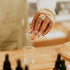Hand with intricate nail art holding a small glass container over a larger glass on a wooden surface with bottles in the foreground.