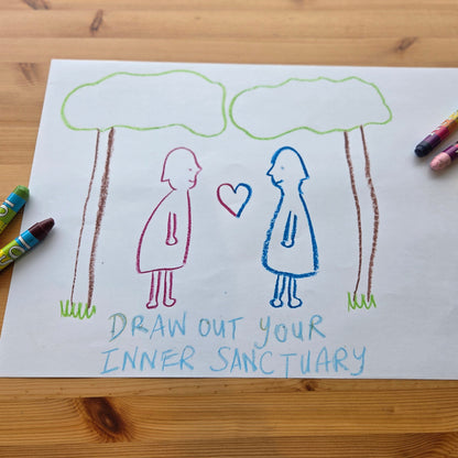 A drawing of two people under clouds with a heart, surrounded by crayons on a wooden surface.