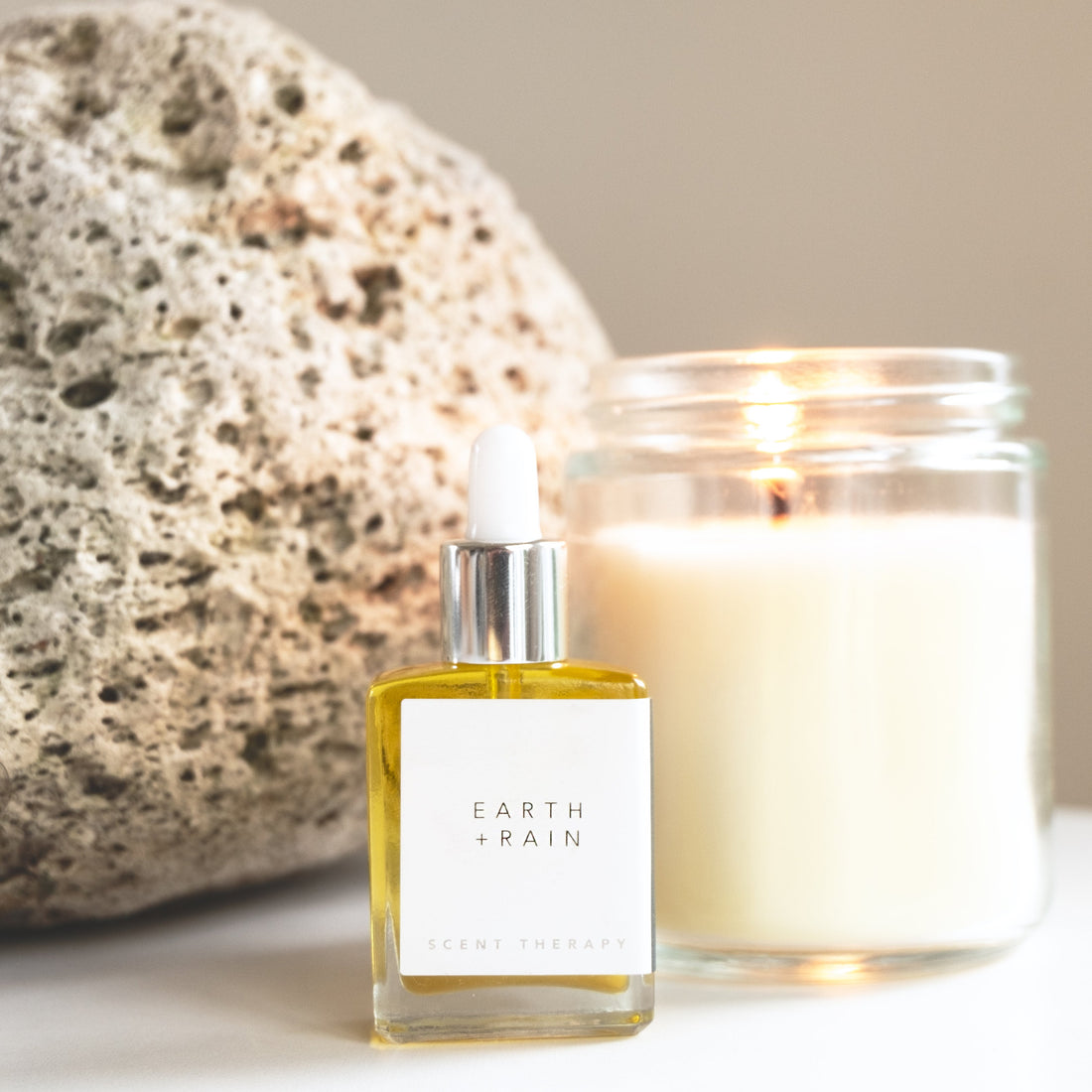 A square glass dropper bottle with the Earth + Rain scent therapy blend, with candle and pumice in the background. 