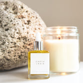 A square glass dropper bottle with the Earth + Rain scent therapy blend, with candle and pumice in the background. 