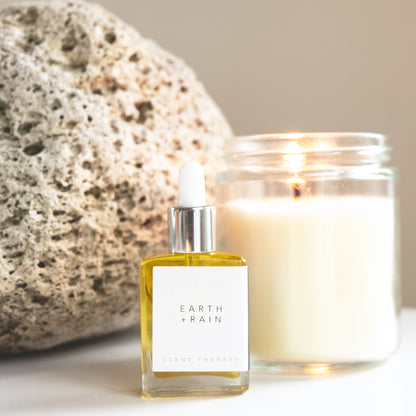 A square glass dropper bottle with the Earth + Rain scent therapy blend, with candle and pumice in the background. 
