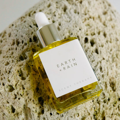 A square glass dropper bottle of the Earth + Rain scent therapy blend lying on a large pumice rock. 