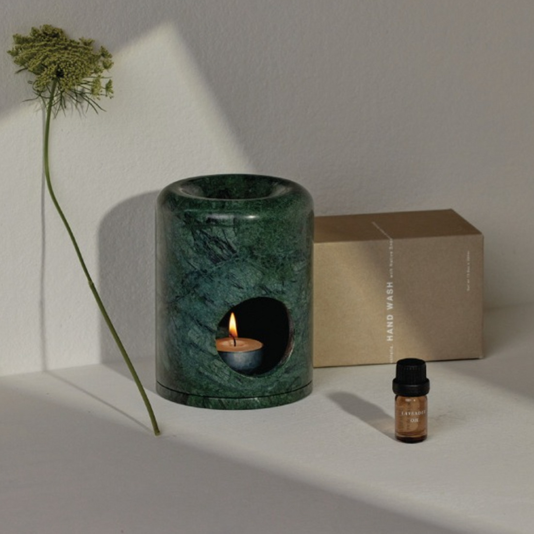 Dark green marble oil burner with lit tea light candle and oil displayed on white surface with a soft sunlight and wild carrot flower. 