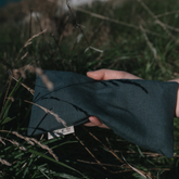 A hand holding a kale coloured linen eye pillow among some dark grasses. 