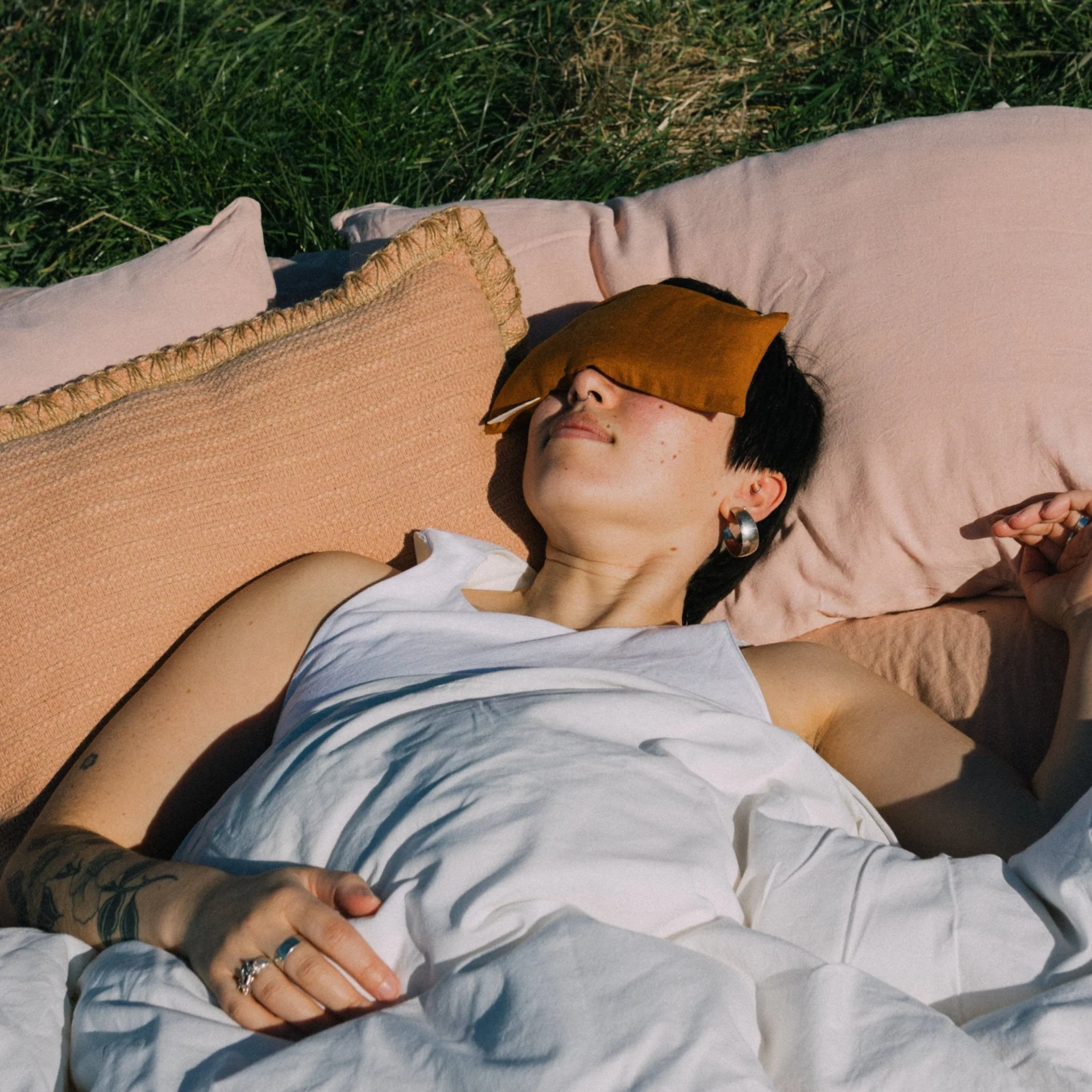 A woman resting on a duvet on the grass in the sunshine with a clay coloured linen eye pillow over her eyes. 