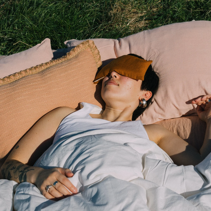 A woman resting on a duvet on the grass in the sunshine with a clay coloured linen eye pillow over her eyes. 