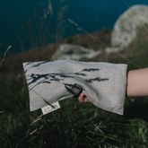 A hand holding a sand coloured linen eye pillow among some dark grasses. 