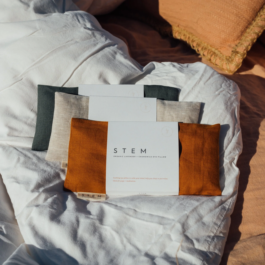 Three linen eye pillows on a duvet in the sunshine, one clay, one sand and one kale. 