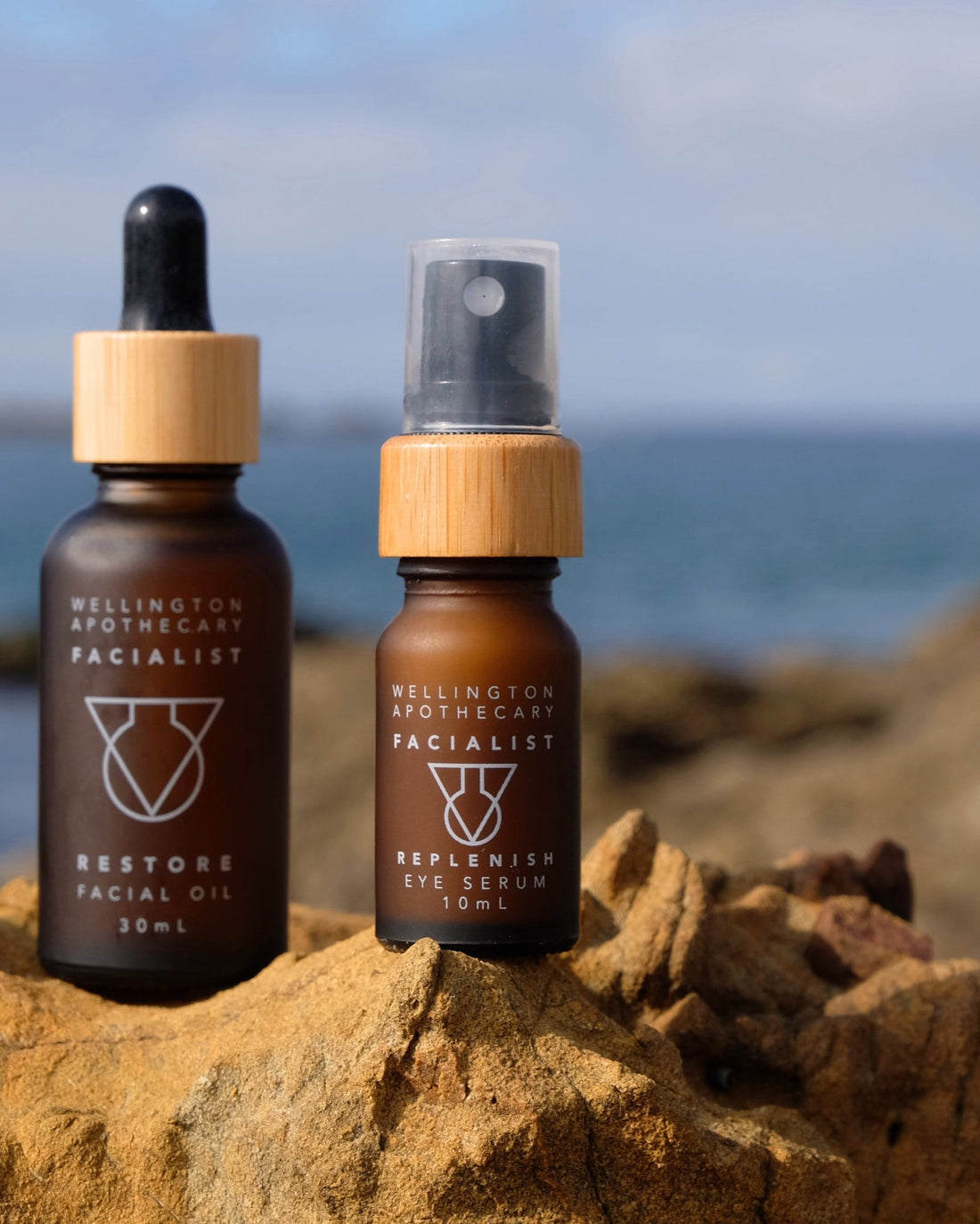 A frosted amber glass bottle of Replenish Eye Serum and a bottle of Restore Facial Oil standing on the south coast rocks in Wellington.