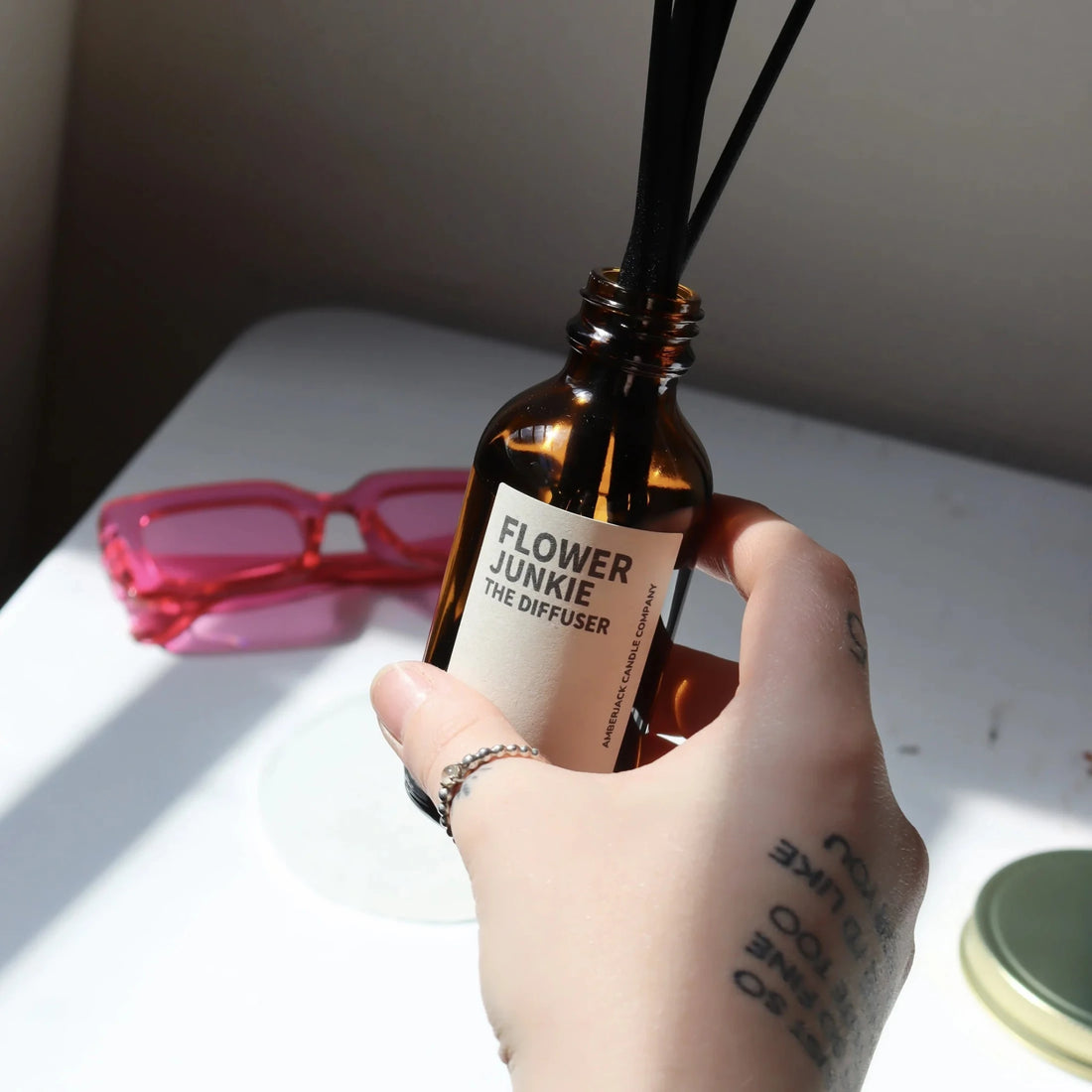 A bottle of Flower Junkie reed diffuser held in hand near a desk with bright pink glasses. 