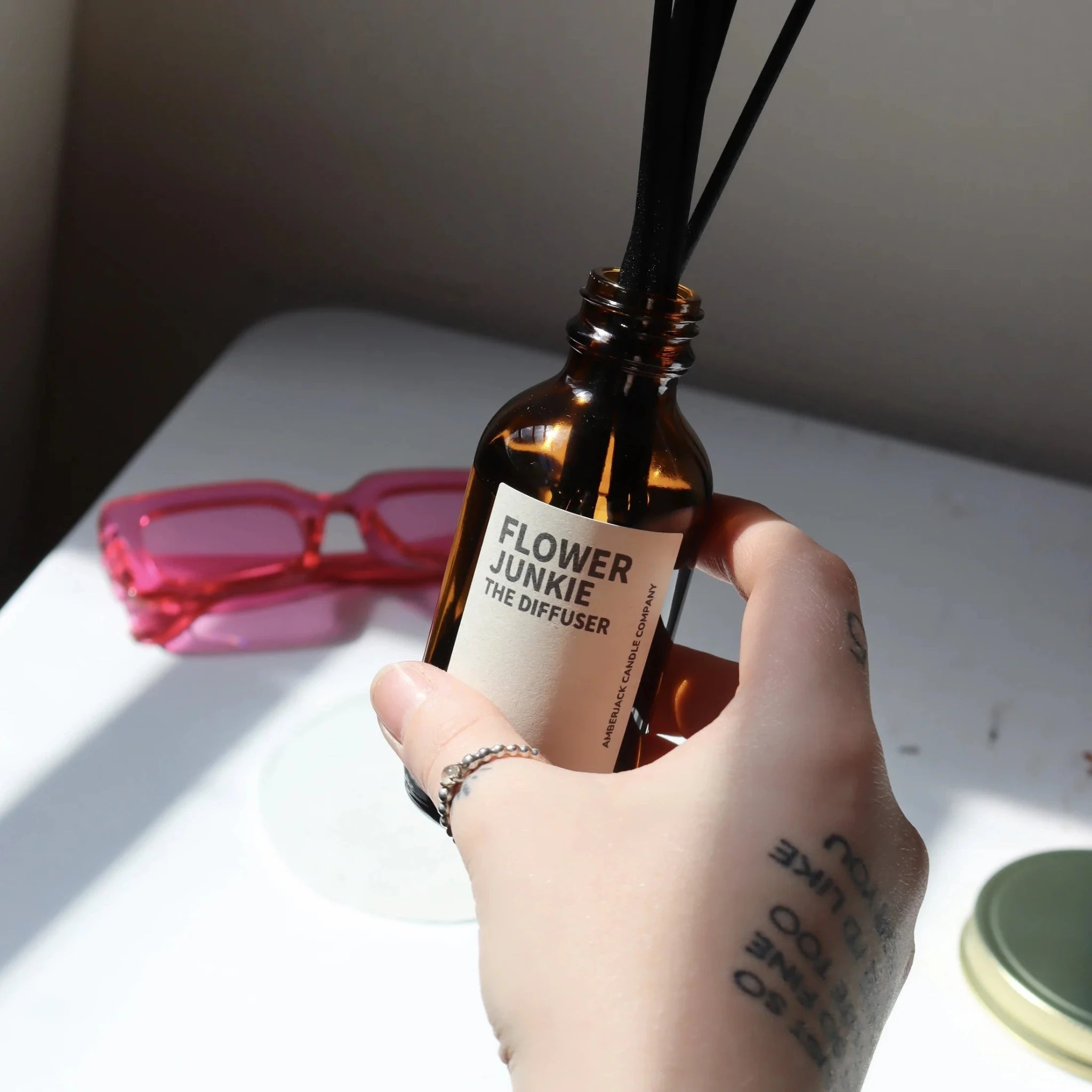A bottle of Flower Junkie reed diffuser held in hand near a desk with bright pink glasses. 