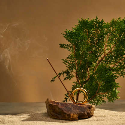 Hinoki incense burner in brass holder a top a rock and sand with conifer branch behind.