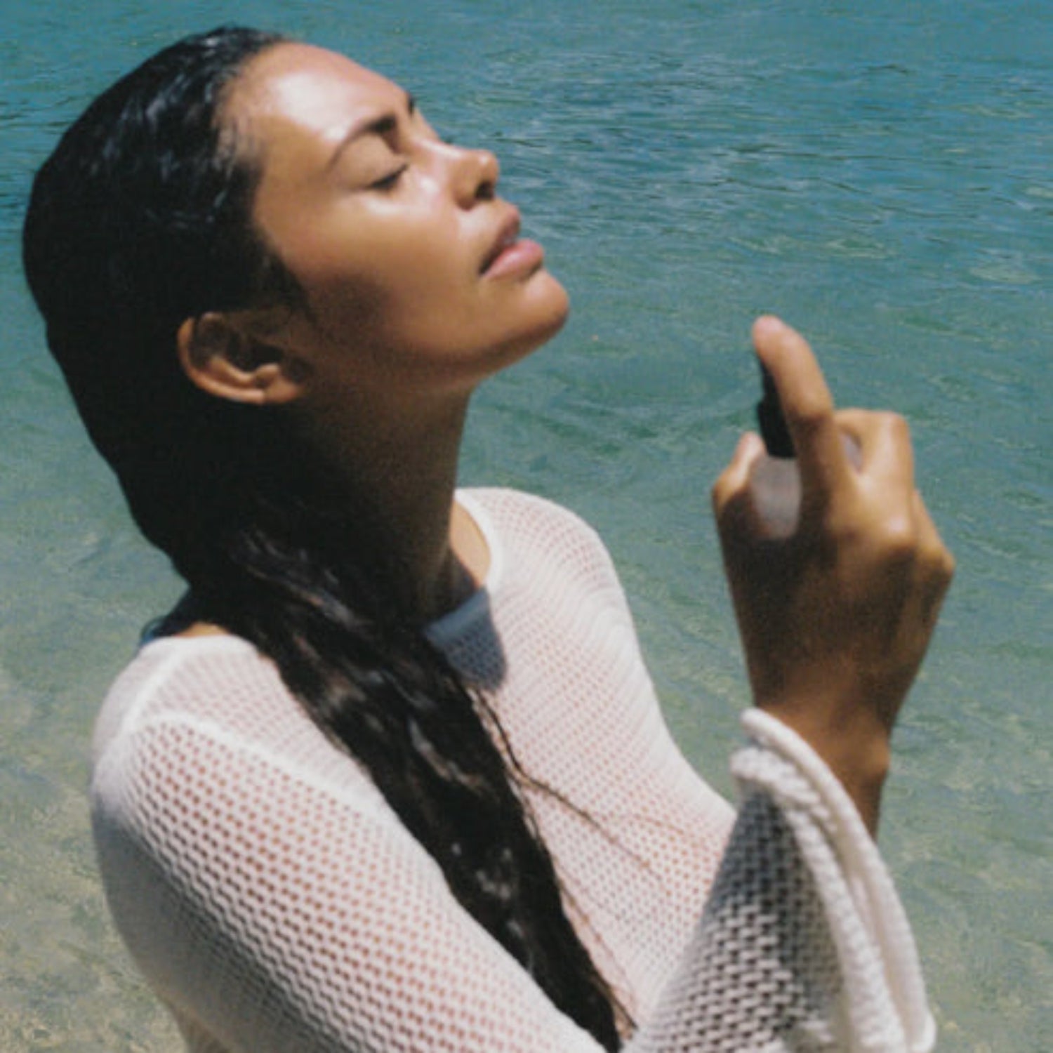 Woman standing by a body of water, wearing a light-colored mesh top.