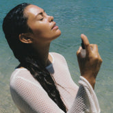 Woman standing by a body of water, wearing a light-colored mesh top.