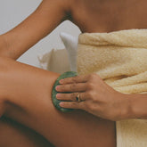 Person using a green jade stone on their leg with a towel draped over them, against a neutral background.