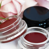 Close up of naturally tinted rose hued lip colours in clear glass pottles, surrounded by rose petals. 