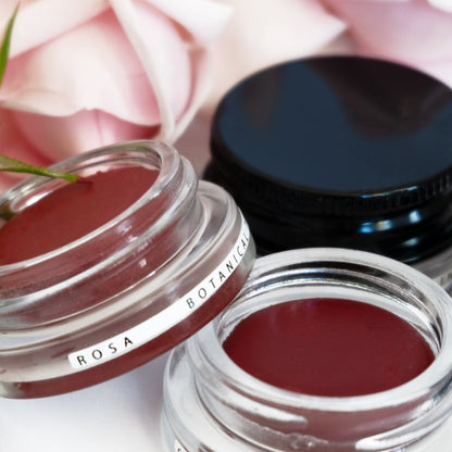 Close up of naturally tinted rose hued lip colours in clear glass pottles, surrounded by rose petals. 