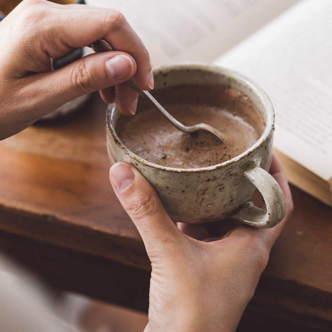 Person holding a ceramic mug with a spoon, sitting on a wooden surface with a book in the background.