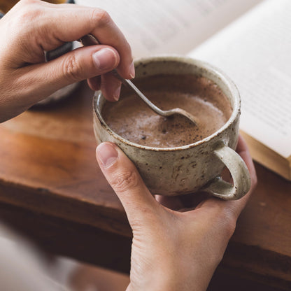 Person holding a ceramic mug with a spoon, sitting on a wooden surface with a book in the background.
