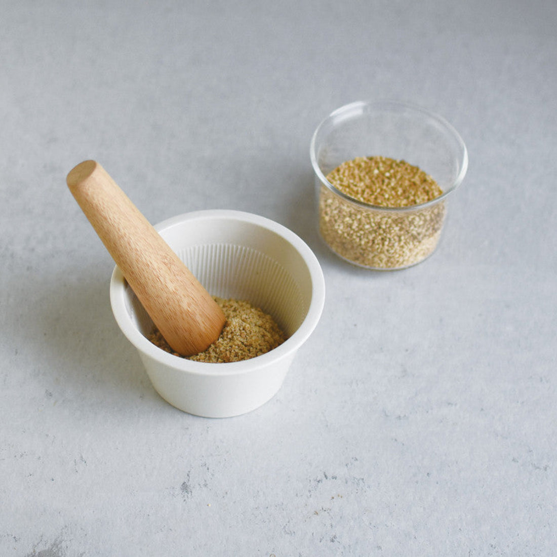 A white ceramic mortar and bamboo pestle on a grey bench with a glass gar of seeds. 