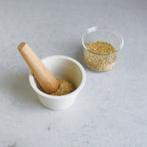 A white ceramic mortar and bamboo pestle on a grey bench with a glass gar of seeds. 