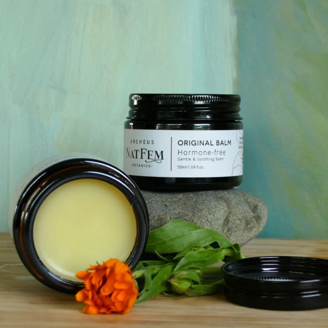 NATFEM Original Balm jar with open lid, balm, calendula flower, and stones on a wooden surface with a green blurred background.