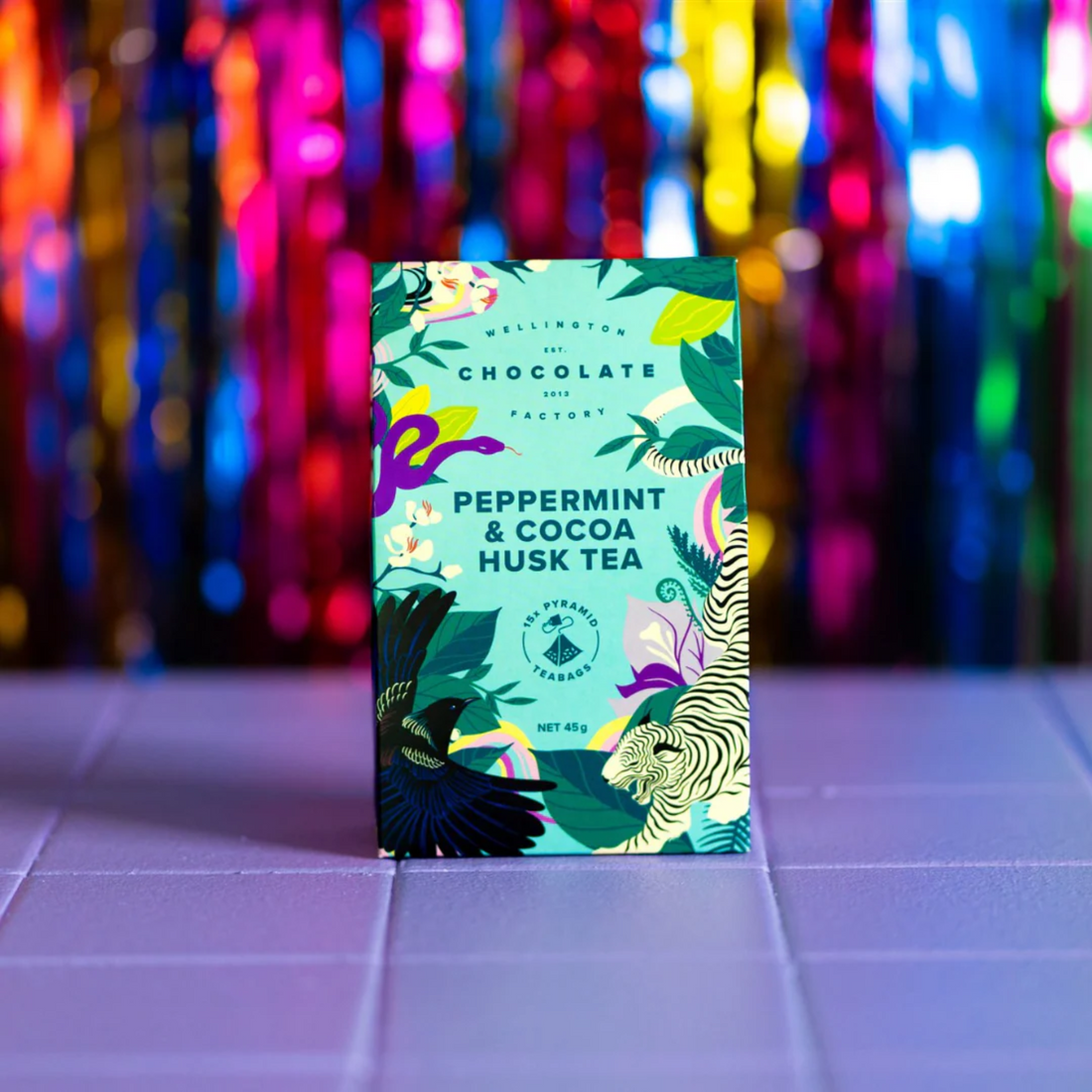 Peppermint cocoa husk tea package with colorful design on a tiled surface with multicoloured lights in the background