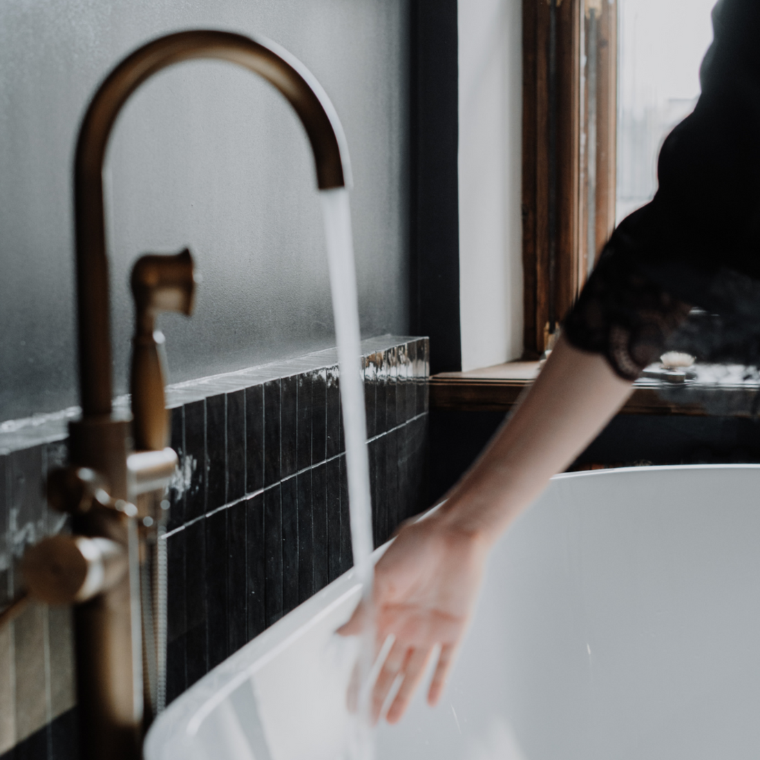 Person washing hands under a running faucet in a bathroom with black tiles and a window.