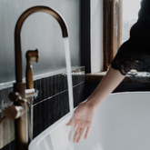 Person washing hands under a running faucet in a bathroom with black tiles and a window.