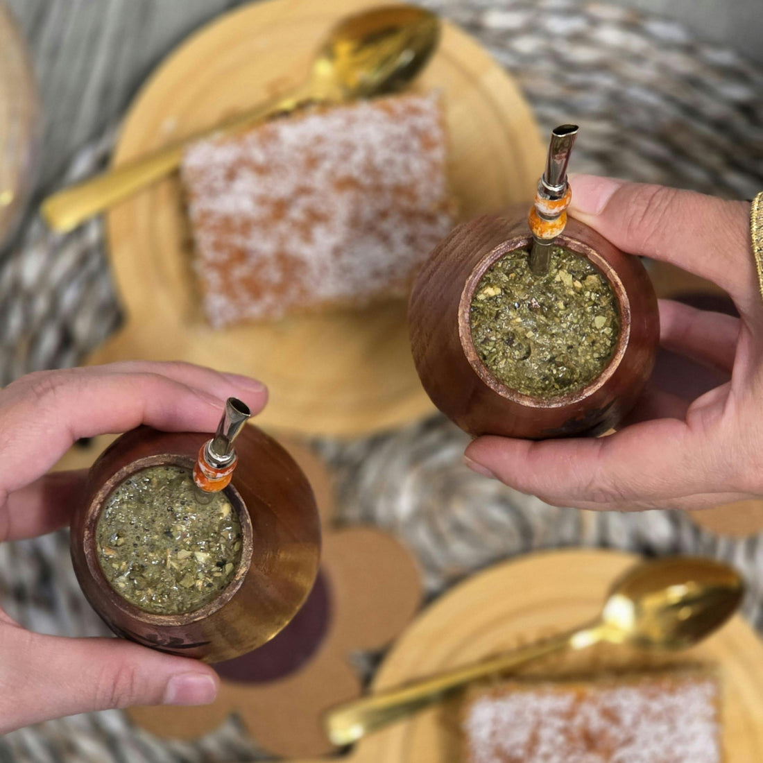 Two hands holding wooden mate gourds filled with green substance, with a plate of sweet food in the background.