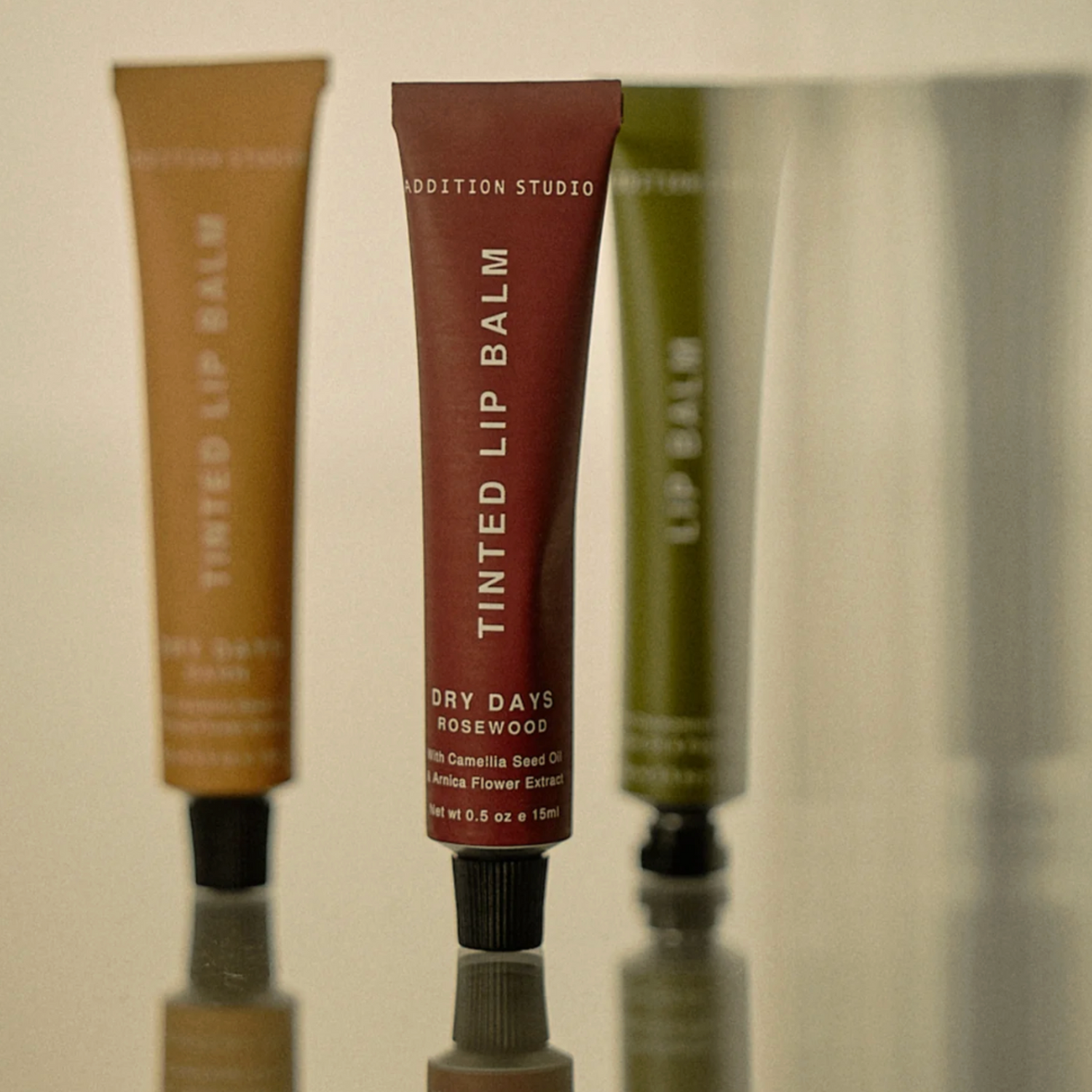 Three tubes of tinted lip balm on a reflective surface with a neutral background