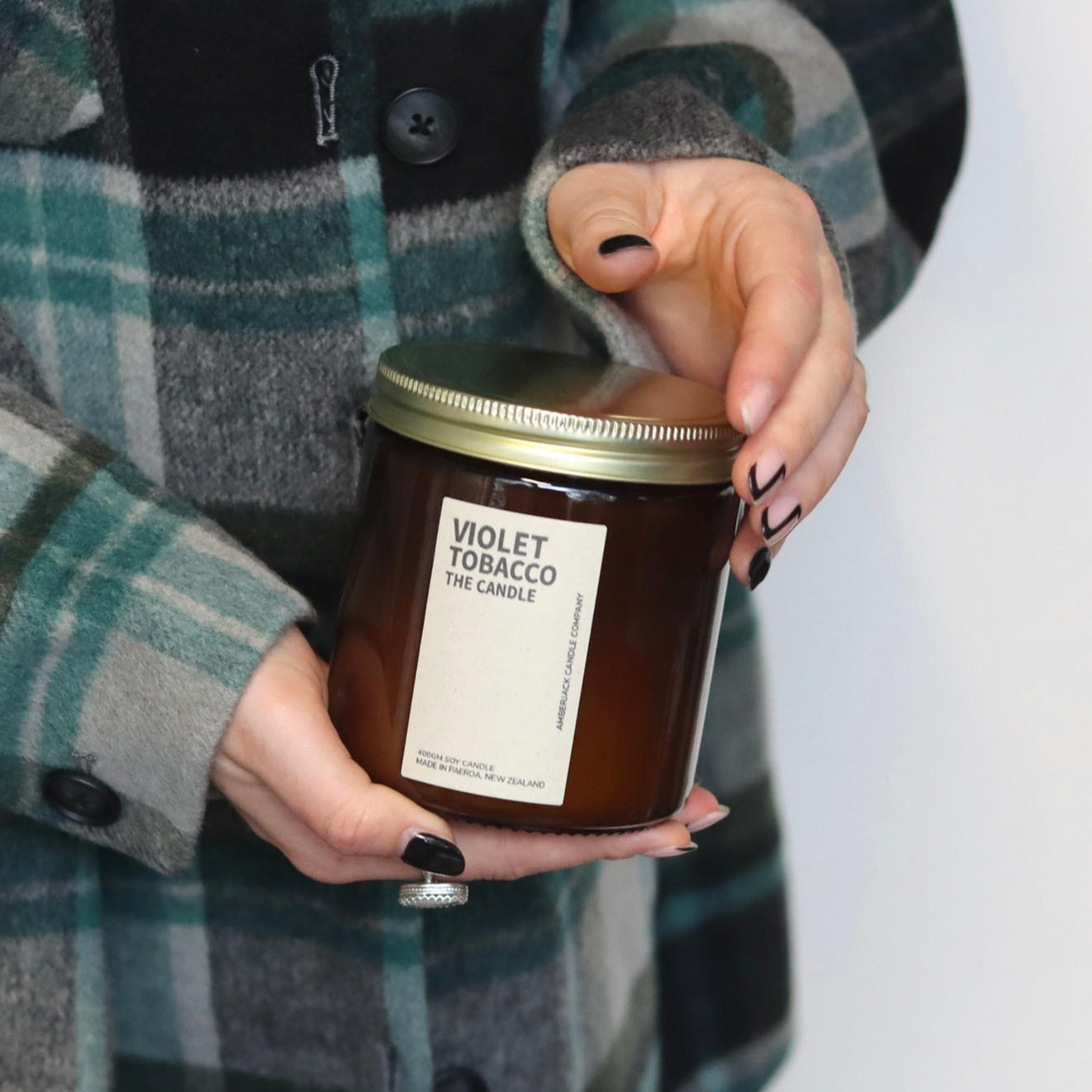 A large amber glass jar with brass lid containing violet tobacco candle held in hands with black nail polish, person wearing teal and grey tartan coat. 