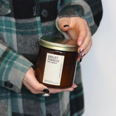A large amber glass jar with brass lid containing violet tobacco candle held in hands with black nail polish, person wearing teal and grey tartan coat. 