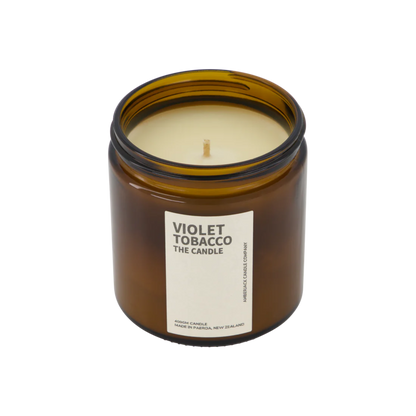 A large amber glass jar with brass lid containing violet tobacco candle on white background. 