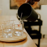 Person pouring liquid candle wax from a black pitcher into glasses on a wooden tray.