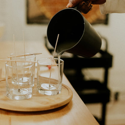 Person pouring liquid candle wax from a black pitcher into glasses on a wooden tray.