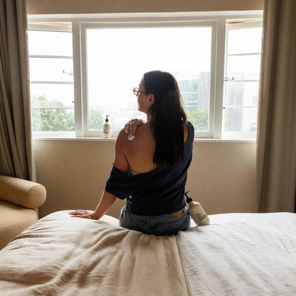 Woman sitting on a bed looking out a window with a bottle of lotion nearby.