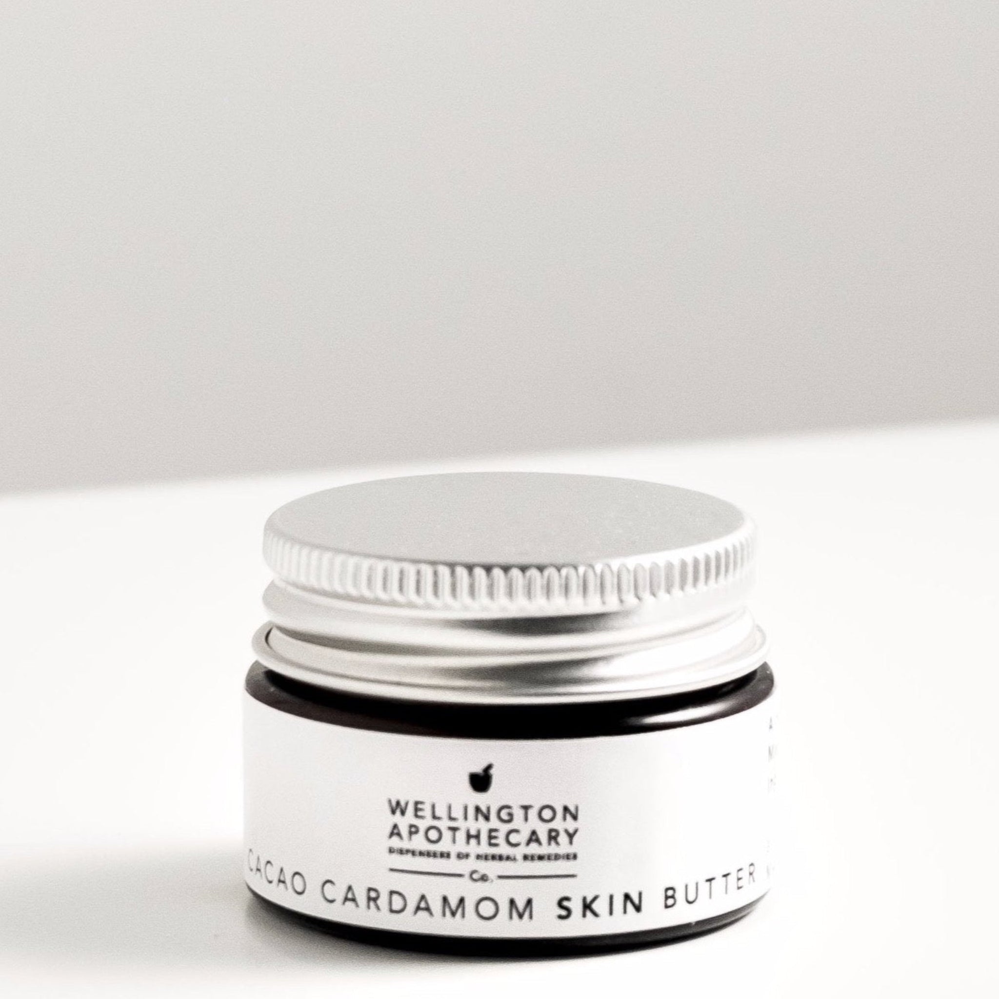 Small amber glass jar of cacao and cardamom lip balm on white bench. 