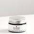 Small amber glass jar of cacao and cardamom lip balm on white bench. 