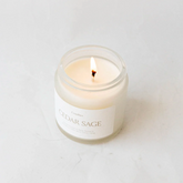 Clear glass jar containing cedar sage candle alight on white background. 
