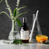 Spray bottle of Holistic Hair scalp spray on bench with orange, rosemary, neroli and conical flasks. 