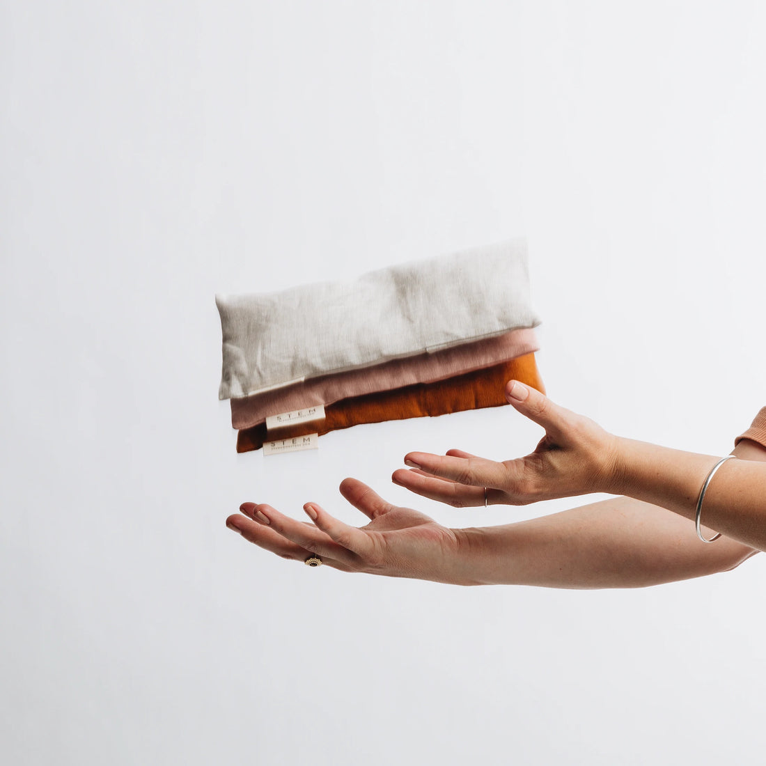 Three linen eye pillows thrown in the air, one sand, one petal and one clay. 
