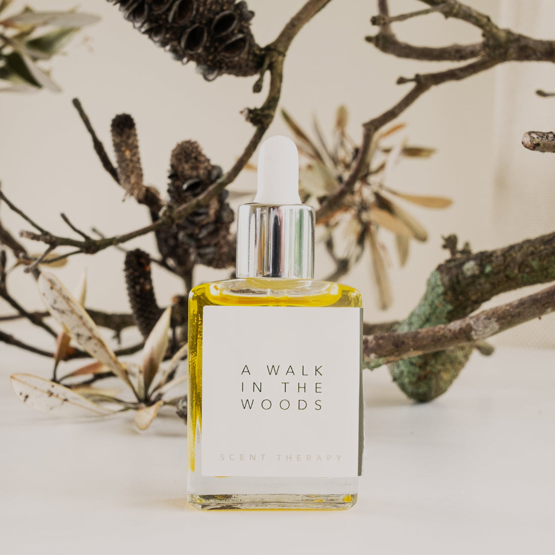 A square glass bottle with dropper top containing the scent therapy blend, a walk in the woods, with old lichen covered twigs and branches in the background.