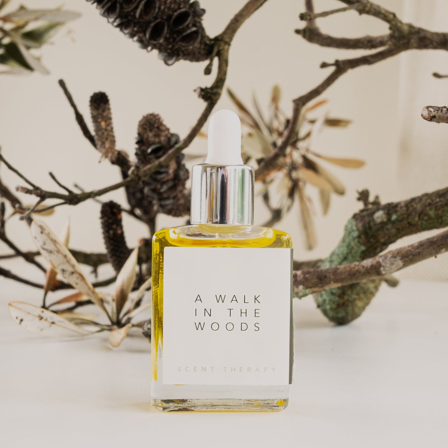 A square glass bottle with dropper top containing the scent therapy blend, a walk in the woods, with old lichen covered twigs and branches in the background.