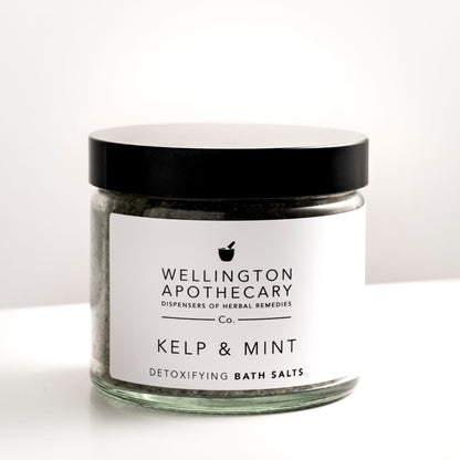 Large clear glass glass filled with kelp and mint bath salts on white bench. 