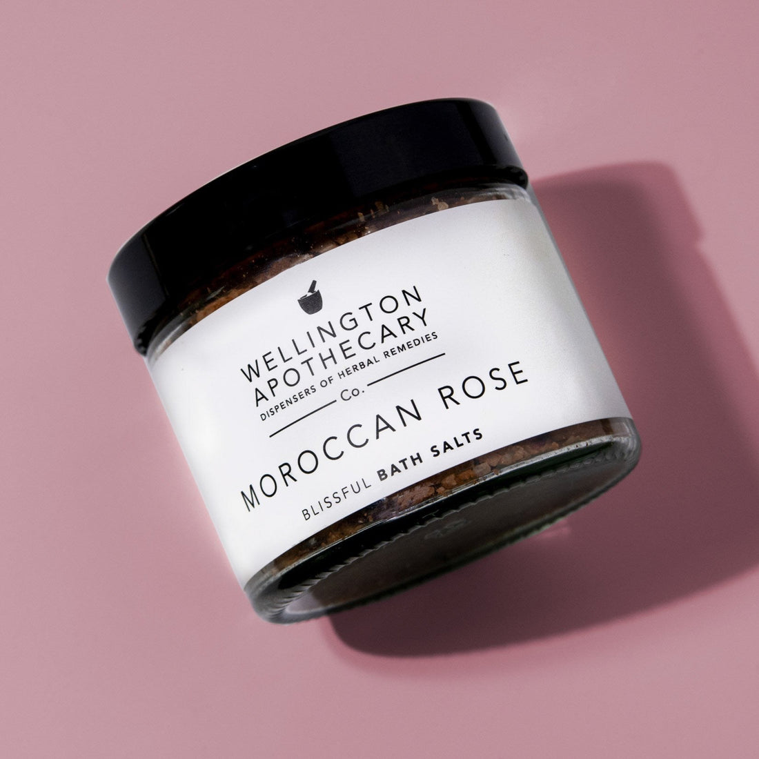 Glass jar filled with bath salts lying on a dusky rose pink surface.